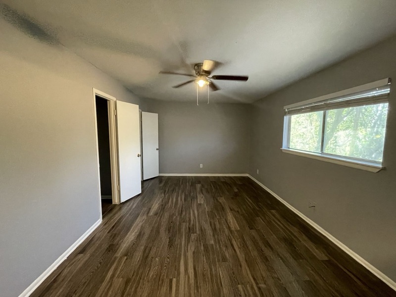 An empty La Bella living room with a ceiling fan, plank floor and daylight from a side window.